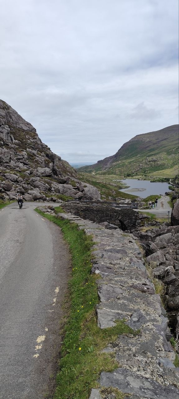 Carretera del Gap of Dunloe flanqueada por muros de piedra, con un puente de arco de piedra sobre el arroyo y una bici pasando
