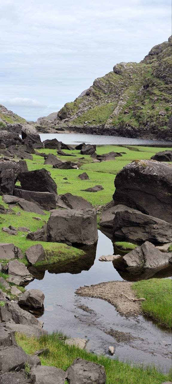 Valle glaciar del Gap of Dunloe: un arroyo serpentea entre bloques de piedra oscuros hacia un lago, entre laderas empinadas