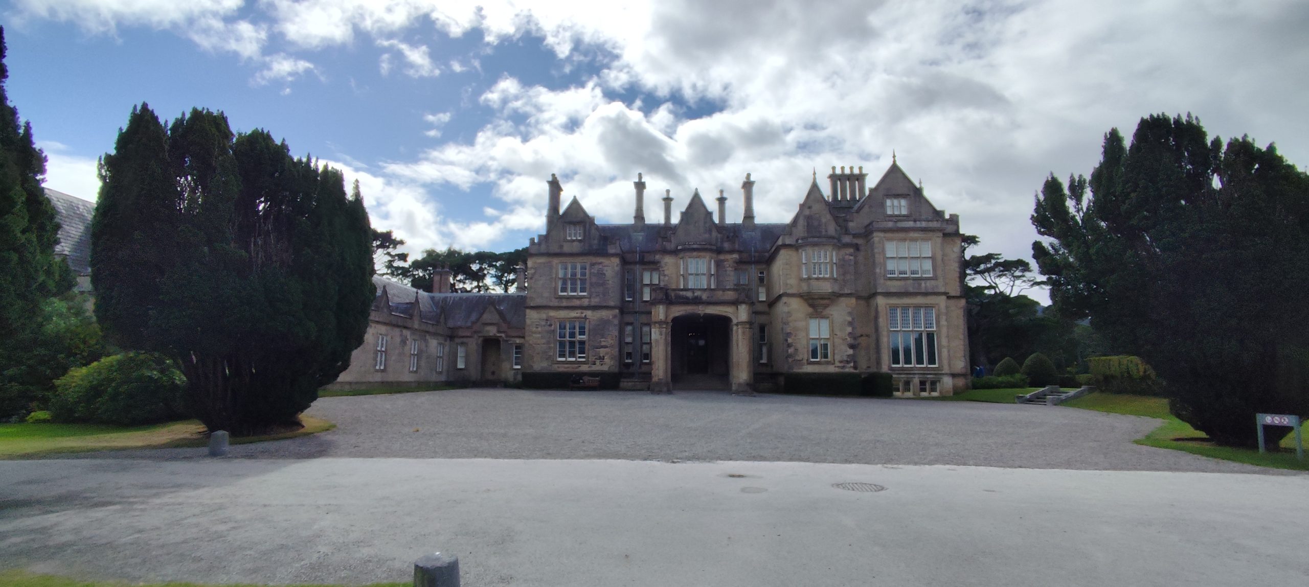 Fachada de Muckross House, mansión de piedra de estilo Tudor con tejados a dos aguas y numerosas chimeneas, rodeada de árboles altos recortados