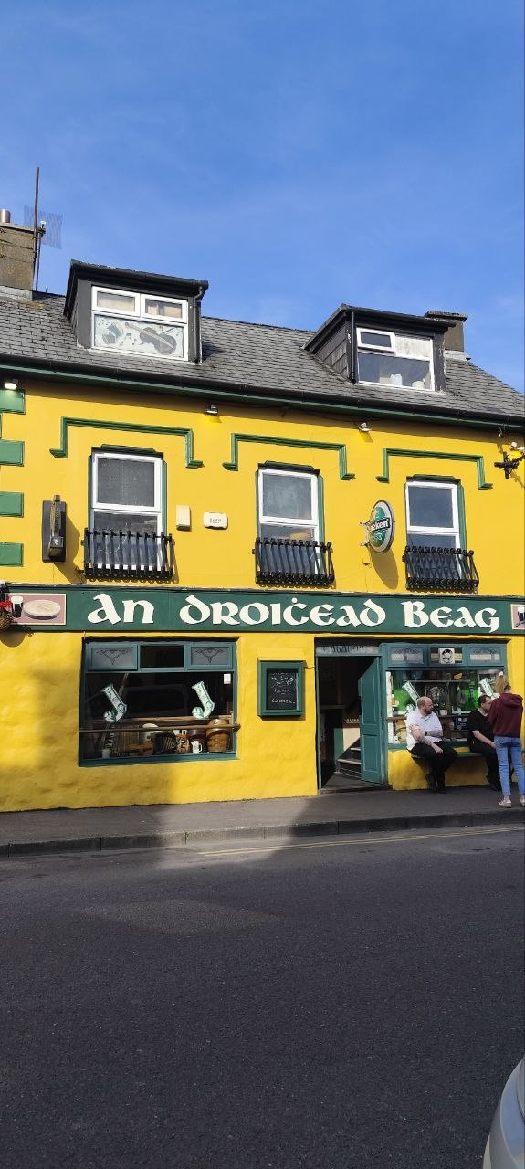 Pub an Droichead Beag en Dingle, edificio amarillo de dos plantas con detalles verdes y terraza con gente sentada