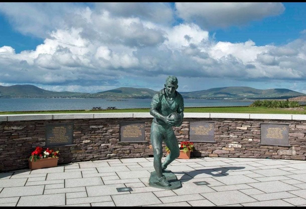 Monumento de bronce a un jugador de rugby en Waterville, con placas conmemorativas y macetas de flores